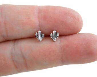 May include: A pair of small, silver-colored cactus stud earrings. Each earring features a detailed cactus design with vertical lines and small side arms. The earrings are shown against a light background.
