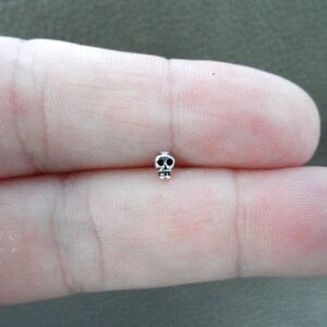 K&ouml;nnte beinhalten: Ein kleiner, silberfarbener Totenkopf-Anh&auml;nger. Der Totenkopf hat detaillierte Merkmale, einschlie&szlig;lich Augenh&ouml;hlen und Kieferlinie. Der Anh&auml;nger ist klein und misst etwa 0,6 cm in der H&ouml;he. Der Anh&auml;nger ist wahrscheinlich aus Metall und hat eine polierte Oberfl&auml;che.