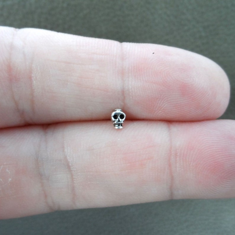 K&ouml;nnte beinhalten: Ein kleiner, silberfarbener Totenkopf-Anh&auml;nger. Der Totenkopf hat detaillierte Merkmale, einschlie&szlig;lich Augenh&ouml;hlen und Kieferlinie. Der Anh&auml;nger ist klein und misst etwa 0,6 cm in der H&ouml;he. Der Anh&auml;nger ist wahrscheinlich aus Metall und hat eine polierte Oberfl&auml;che.