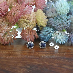 Pu&ograve; includere: Orecchini a bottone in argento con una pietra centrale blu scuro macchiettata e un bordo decorativo. Sono visibili anche due chiusure per orecchini. Gli orecchini sono esposti su una superficie di legno con accenti floreali colorati sullo sfondo.