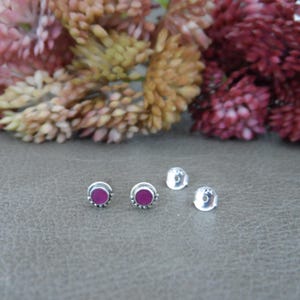 May include: A pair of silver stud earrings with a small, round, dark pink stone in the centre of each earring.
