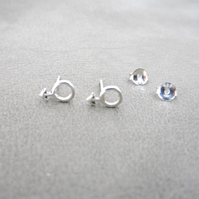 May include: A pair of silver stud earrings featuring a male symbol design. The earrings are marked with "925" on the back.