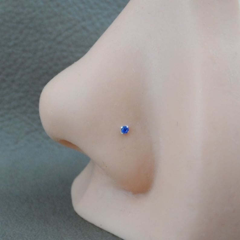 Op de afbeelding: Een close-up van een zilveren neuspiercing met een kleine, blauwe edelsteen.