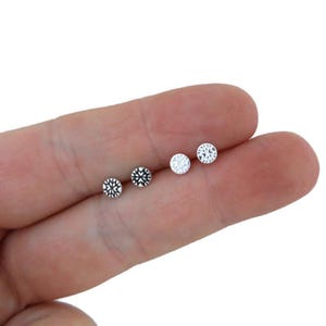 Può includere: Quattro piccoli orecchini a bottone. Due sono argentati con un motivo a fiocco di neve e due sono trasparenti con un motivo sfaccettato. Gli orecchini sono piccoli e rotondi. Gli orecchini sono mostrati su una mano.