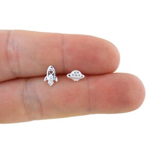 May include: A pair of silver stud earrings featuring a rocket and a planet design. The rocket has a heart-shaped window and the planet has a ring around it. Both earrings are decorated with small, clear cubic zirconia stones.