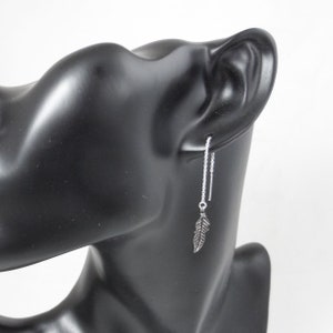 May include: Silver feather dangle earrings with a delicate chain. The earrings are shown on a black mannequin head.