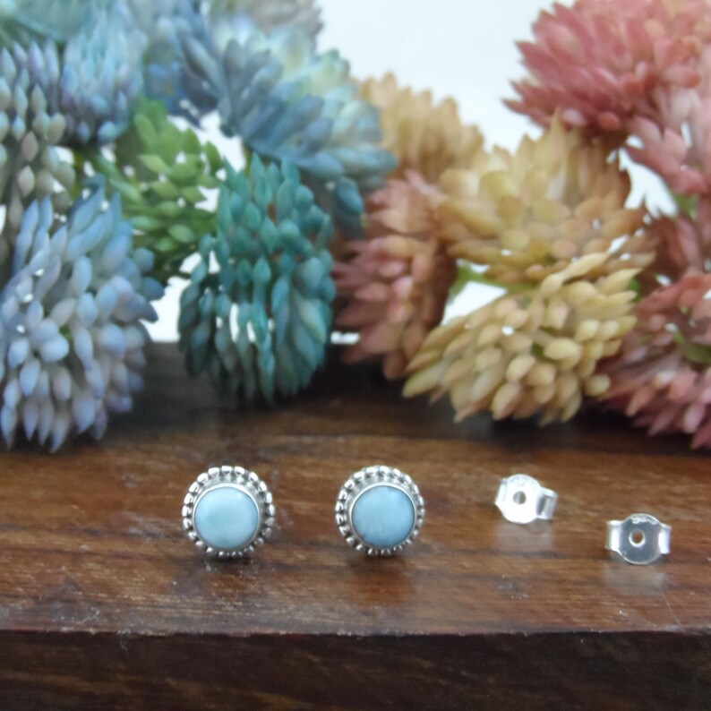 May include: A pair of silver stud earrings with a light blue gemstone in the centre. The earrings have a beaded border around the gemstone.