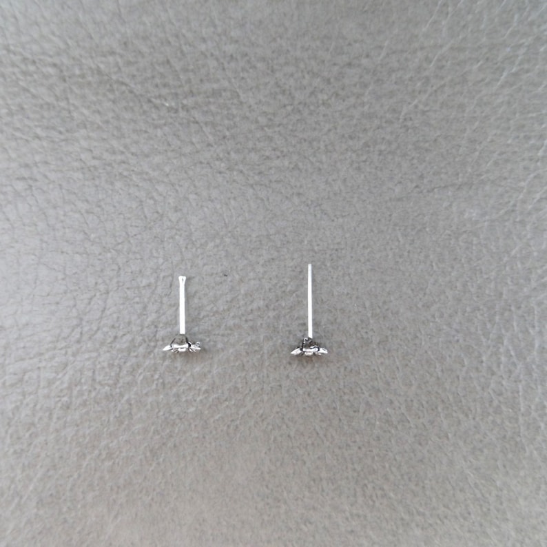 K&ouml;nnte beinhalten: Zwei kleine, silberfarbene Ohrstecker. Jeder Ohrring hat einen d&uuml;nnen Stift und eine dekorative Ansammlung kleiner, gl&auml;nzender Elemente an der Basis. Die Ohrringe liegen auf einer strukturierten, grauen Oberfl&auml;che.