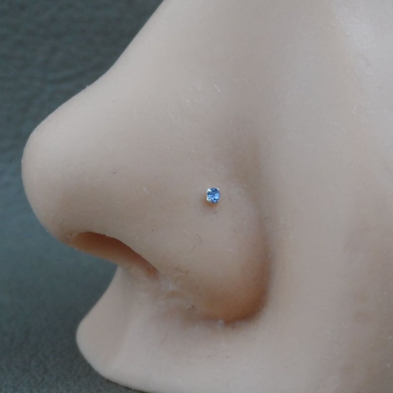 May include: A close-up of a light blue gemstone nose ring stud. The stud is set in a silver nose ring.