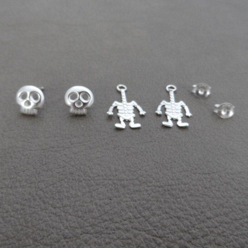 May include: A set of silver earring findings featuring a skull and skeleton design. The set includes two skull earrings, two skeleton charms, and two earring backs.