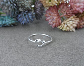 Könnte beinhalten: Ein silberner Knotenring mit einem schlichten Design. Der Ring ist dünn und zart, und der Knoten ist glatt und poliert.