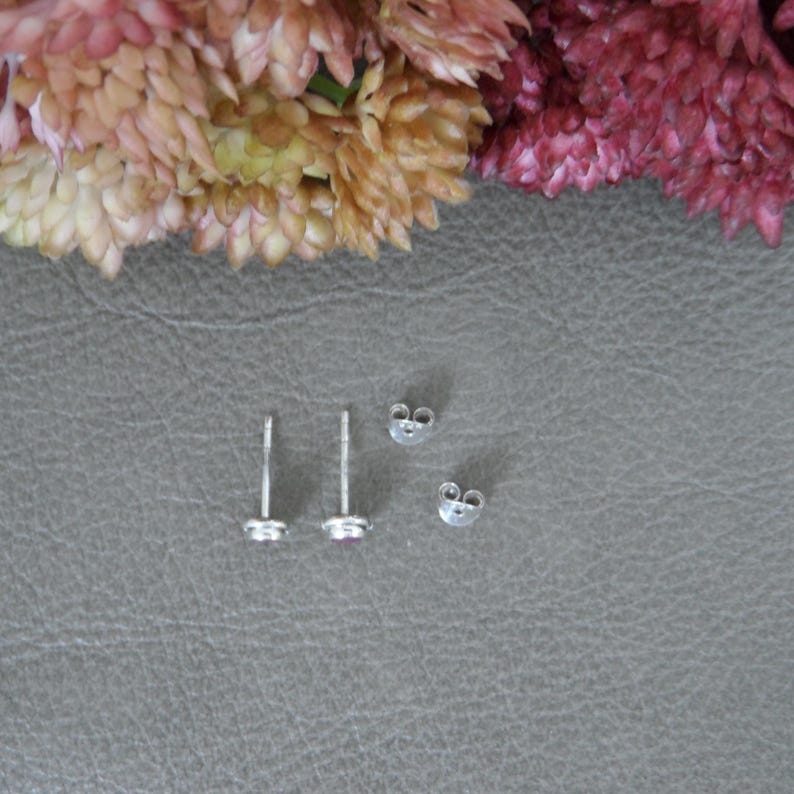 May include: A pair of silver stud earrings with small, clear gemstones. The earrings are shown on a grey surface with two sets of earring backs.