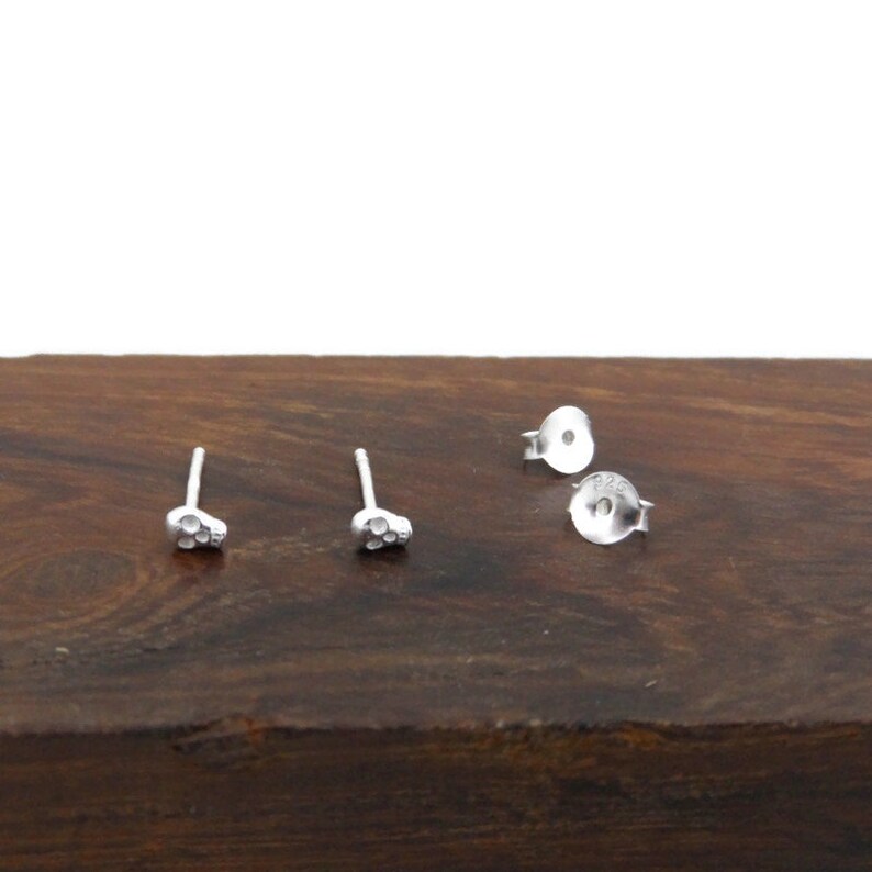 May include: A pair of silver skull stud earrings. The earrings are small and delicate, and they have a textured surface.