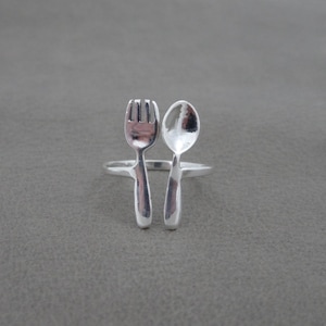 May include: A silver ring with a fork and spoon design. The fork and spoon are connected at the base and form a ring.