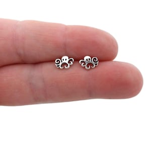 May include: A pair of small, silver-colored octopus stud earrings. Each earring features a stylized octopus design with a round head and curling tentacles. The earrings are shown against a light background.