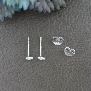 May include: A pair of silver stud earrings with a small, textured design. The earrings are shown on a gray surface.
