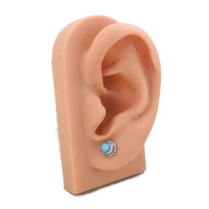 May include: A close-up of a silver earring with a light blue gemstone set in a silver bezel. The earring is on a model ear.