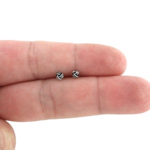 May include: A pair of silver knot stud earrings. The earrings are small and delicate, and they have a simple, elegant design.