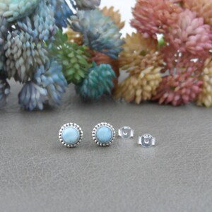 May include: A pair of silver stud earrings with a light blue gemstone set in a beaded bezel. The earrings are shown on a grey surface with a floral background.