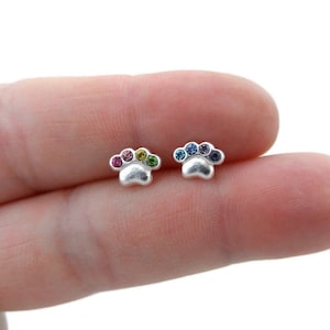 May include: A pair of silver-colored paw print stud earrings. Each earring features a paw print design with five colorful gemstones. The gemstones are arranged in a rainbow pattern on one earring and blue hues on the other.
