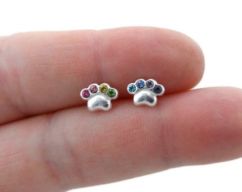 May include: A pair of silver-colored paw print stud earrings. Each earring features a paw print design with five colorful gemstones. The gemstones are arranged in a rainbow pattern on one earring and blue hues on the other.