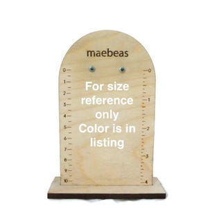 May include: A wooden earring display stand with a ruler on the side. The stand has two green gemstone earrings on it. The text "maebeas" is at the top of the stand. The text "For size reference only" and "Color is in listing" is printed on the stand.