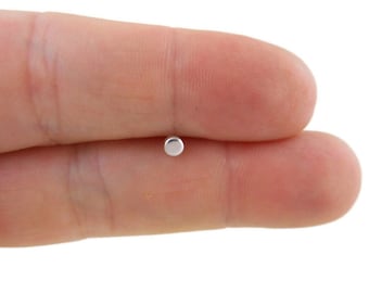 May include: A small, silver-colored, round object, possibly a button or a bead, is held between two fingers. The object is reflective and appears to be made of metal. The background is a blurred, neutral color.