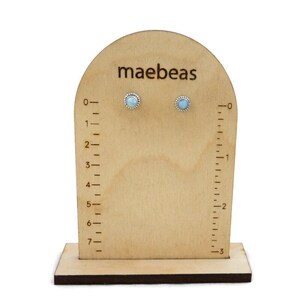 May include: A pair of light blue gemstone stud earrings on a wooden display stand with a ruler. The stand is labelled "maebeas".