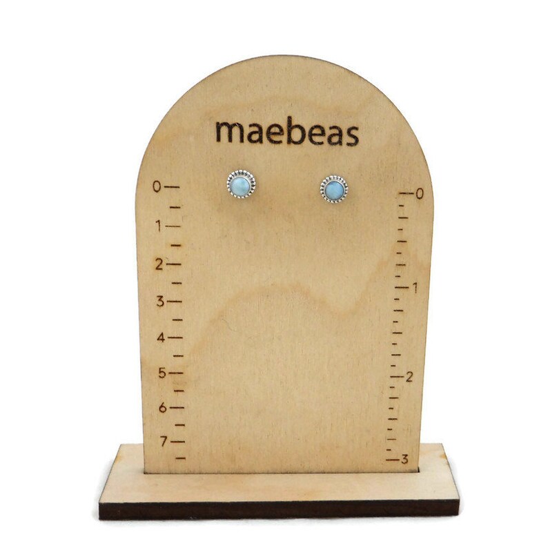 May include: A pair of light blue gemstone stud earrings on a wooden display stand with a ruler. The stand is labelled "maebeas".