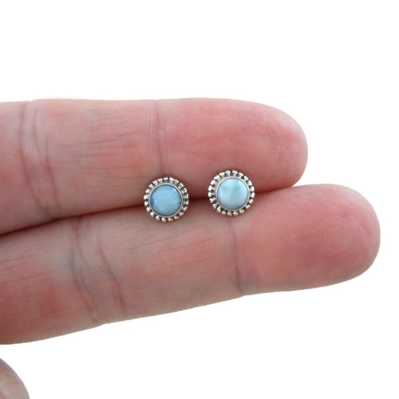 May include: A pair of silver stud earrings with a light blue gemstone in the centre of each earring. The gemstones are surrounded by a silver beaded border.