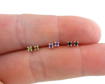 Può includere: Quattro paia di piccoli orecchini a perno d'argento con pietre preziose di diversi colori. Gli orecchini presentano una pietra preziosa verde, viola, nera e nera.
