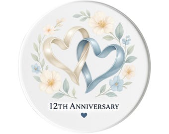 Puede incluir: Un objeto decorativo redondo blanco con dos cintas entrelazadas en forma de corazón en beige y azul. El diseño incluye detalles florales y el texto "12º ANIVERSARIO" con un pequeño símbolo de corazón.