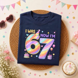 May include: A navy blue t-shirt featuring a colorful graphic with the text "I WAS" and "NOW I'M 67". The design includes a large number 67 in pastel rainbow stripes, a party hat, a birthday cake with candles, balloons, and a gift box.