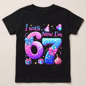 May include: A black short-sleeve t-shirt featuring a colorful graphic with the text I was 6, Now I'm 7. The design includes a pink leopard print number 6 with a blue bow, a blue polka dot number 7 with a pink bow, party hats, balloons, and a cupcake.