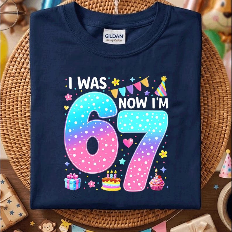 May include: A navy blue t-shirt featuring the white text I WAS NOW I'M 67. The large numbers 6 and 7 have a blue to pink gradient with white polka dots. The design includes colorful party bunting, a party hat, a gift box, and a birthday cake.