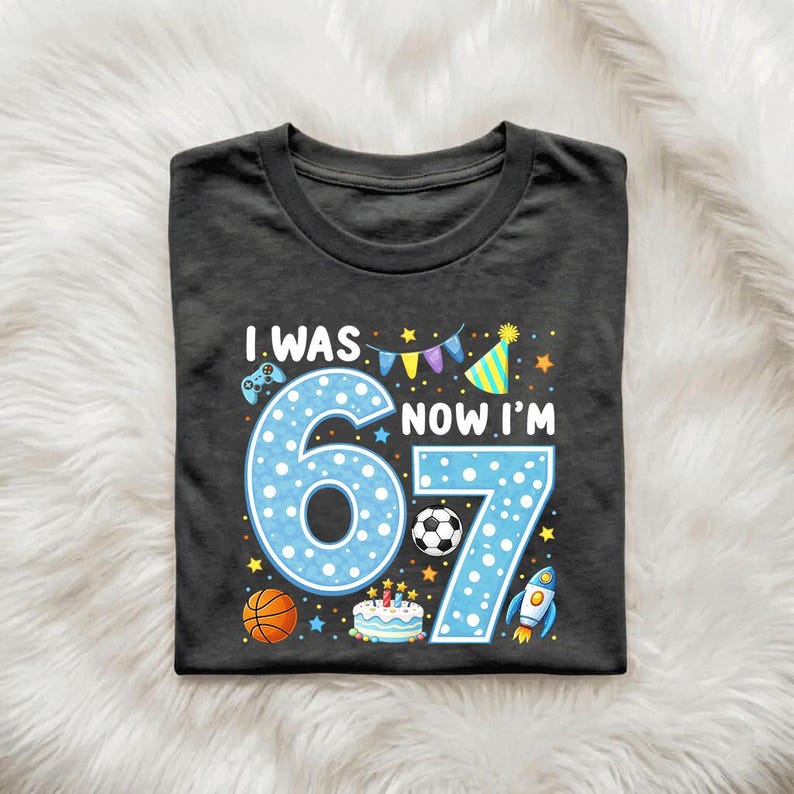 May include: A charcoal gray t-shirt featuring a graphic with the text I WAS 6 NOW I'M 7. The design includes a large light blue polka-dot number 67, a soccer ball, a basketball, a birthday cake, a party hat, a rocket ship, and a game controller.