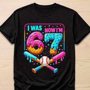 May include: A black short-sleeve t-shirt featuring a graphic with the text I WAS NOW I'M 67. The design includes a pink donut-shaped number 6 and a blue donut-shaped number 7, both with sprinkles, above a baseball and two crossed baseball bats.