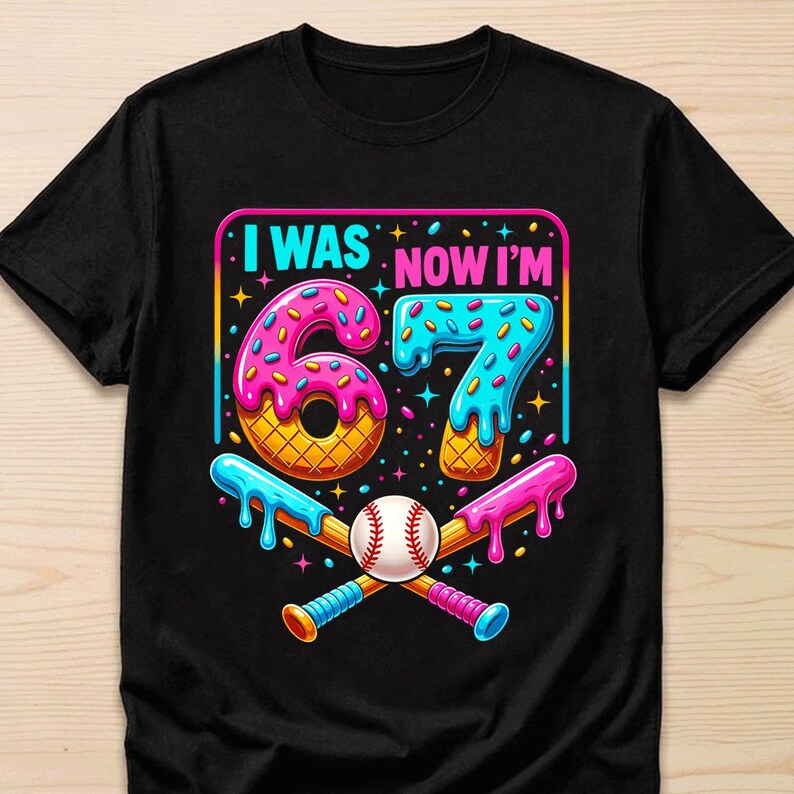 May include: A black short-sleeve t-shirt featuring a graphic with the text I WAS NOW I'M 67. The design includes a pink donut-shaped number 6 and a blue donut-shaped number 7, both with sprinkles, above a baseball and two crossed baseball bats.