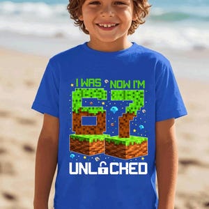 May include: A bright blue short-sleeve t-shirt featuring a graphic print with the text I WAS NOW I'M 67 UNLOCKED. The design includes pixelated block graphics resembling grass and dirt, along with small diamond and star icons in various colors.
