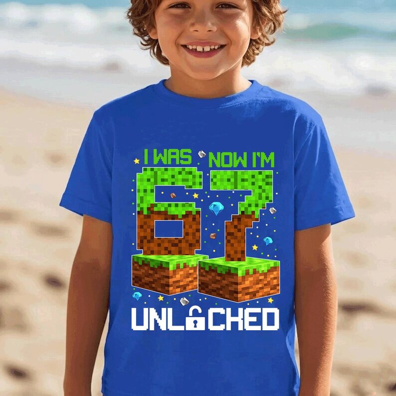 May include: A bright blue short-sleeve t-shirt featuring a graphic print with the text I WAS NOW I'M 67 UNLOCKED. The design includes pixelated block graphics resembling grass and dirt, along with small diamond and star icons in various colors.