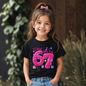 May include: A black short-sleeve t-shirt featuring a graphic with the text I Was Now I'm 67. The numbers 6 and 7 are designed to look like pink frosted donuts with colorful sprinkles, bows, and small party icons like balloons and cupcakes.