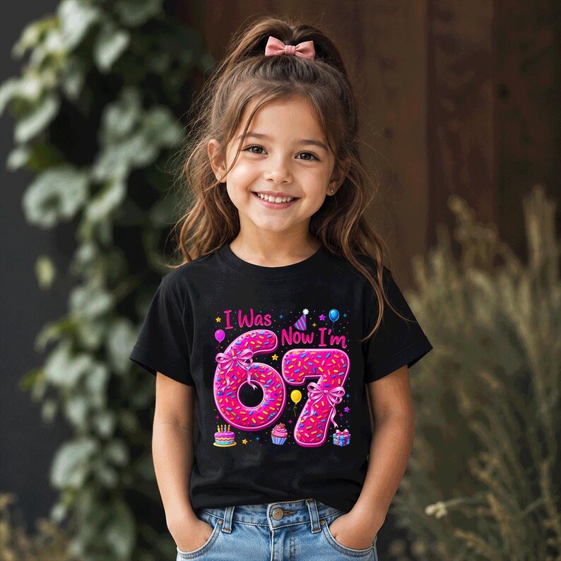 May include: A black short-sleeve t-shirt featuring a graphic with the text I Was Now I'm 67. The numbers 6 and 7 are designed to look like pink frosted donuts with colorful sprinkles, bows, and small party icons like balloons and cupcakes.