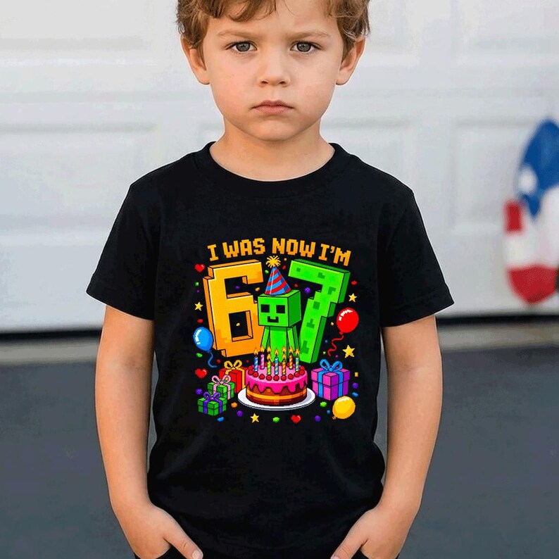 May include: A black short-sleeved t-shirt featuring a colorful graphic with the text I WAS NOW I'M 6 7. The design includes a green pixelated character wearing a party hat, a birthday cake with lit candles, gift boxes, and various balloons.