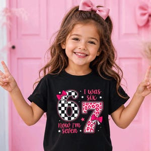 May include: A black short-sleeved t-shirt featuring a graphic with the number 6 in a black and white checkered pattern and the number 7 in a pink leopard print. The text reads I was six now I'm seven, accented with pink bows and stars.