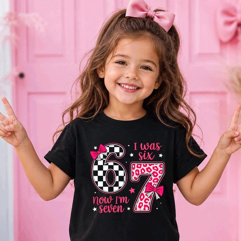 May include: A black short-sleeved t-shirt featuring a graphic with the number 6 in a black and white checkered pattern and the number 7 in a pink leopard print. The text reads I was six now I'm seven, accented with pink bows and stars.
