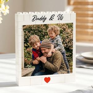 Op de afbeelding: Een witte fotolijst in baksteenstijl met de tekst "Daddy & Us". De lijst bevat een foto van een man en twee kinderen. Onderaan de lijst staat een rood hart. Op de achtergrond staat een vaas met bloemen.