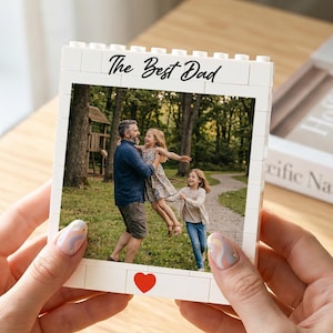 Op de afbeelding: Een witte fotolijst met de tekst "The Best Dad" en een rood hart. De lijst bevat een familiefoto van een vader en twee dochters. De lijst wordt vastgehouden door een persoon met verzorgde nagels.