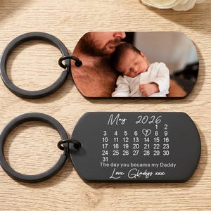 Peut inclure: Deux porte-clés noirs. L'un présente une photo d'un bébé et d'une personne. L'autre porte-clés affiche le mois de mai 2026 avec un calendrier et le texte "The day you became my Daddy Love Gladys xxx."