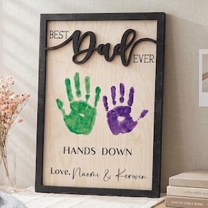 Può includere: Targa in legno incorniciata con la scritta "BEST Dad EVER" in corsivo. La targa presenta impronte di mani verdi e viola, il testo "HANDS DOWN" e "Love, Naomi & Kerwin".