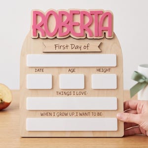 May include: A wooden arch-shaped sign with the name ROBERTA in pink letters at the top. It features the text First Day of, DATE, AGE, HEIGHT, THINGS I LOVE, and WHEN I GROW UP, I WANT TO BE, with blank white rectangular spaces for writing.